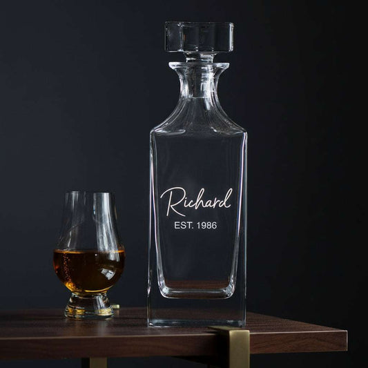 Clear glass decanter with engraved name and year on a wooden table with a blurred light background