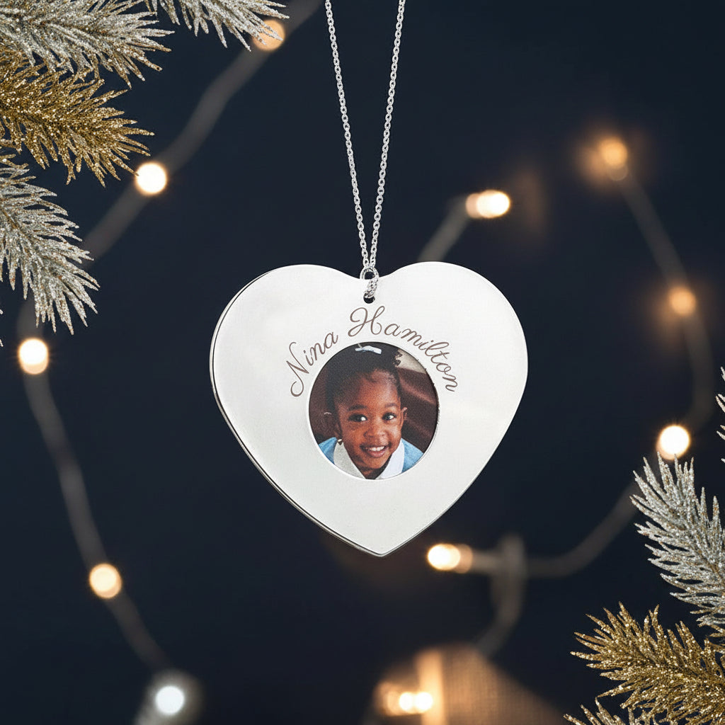 Heart-shaped ornament with a photo of a child and engraved name, hanging on a white decorative branch.
