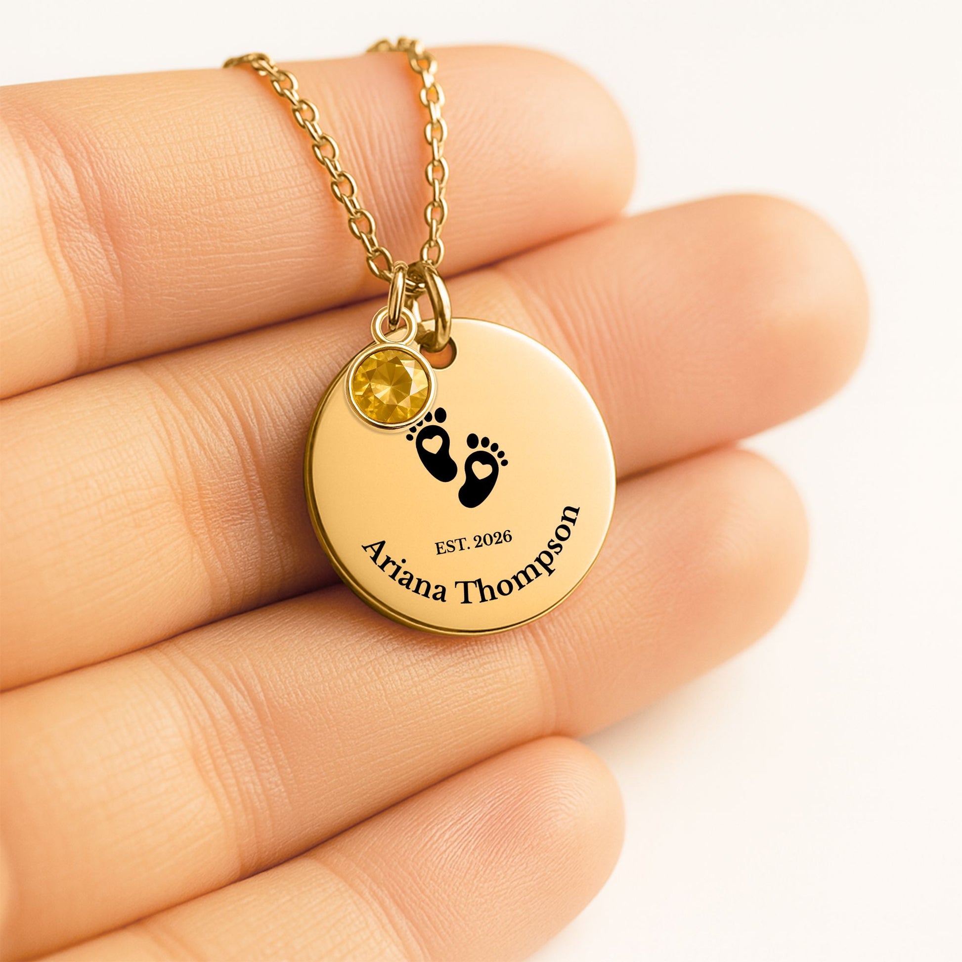 A gold round pendant necklace with black engraved text and baby footprints, held on a person's fingers. The pendant also features a small round yellow gemstone.
