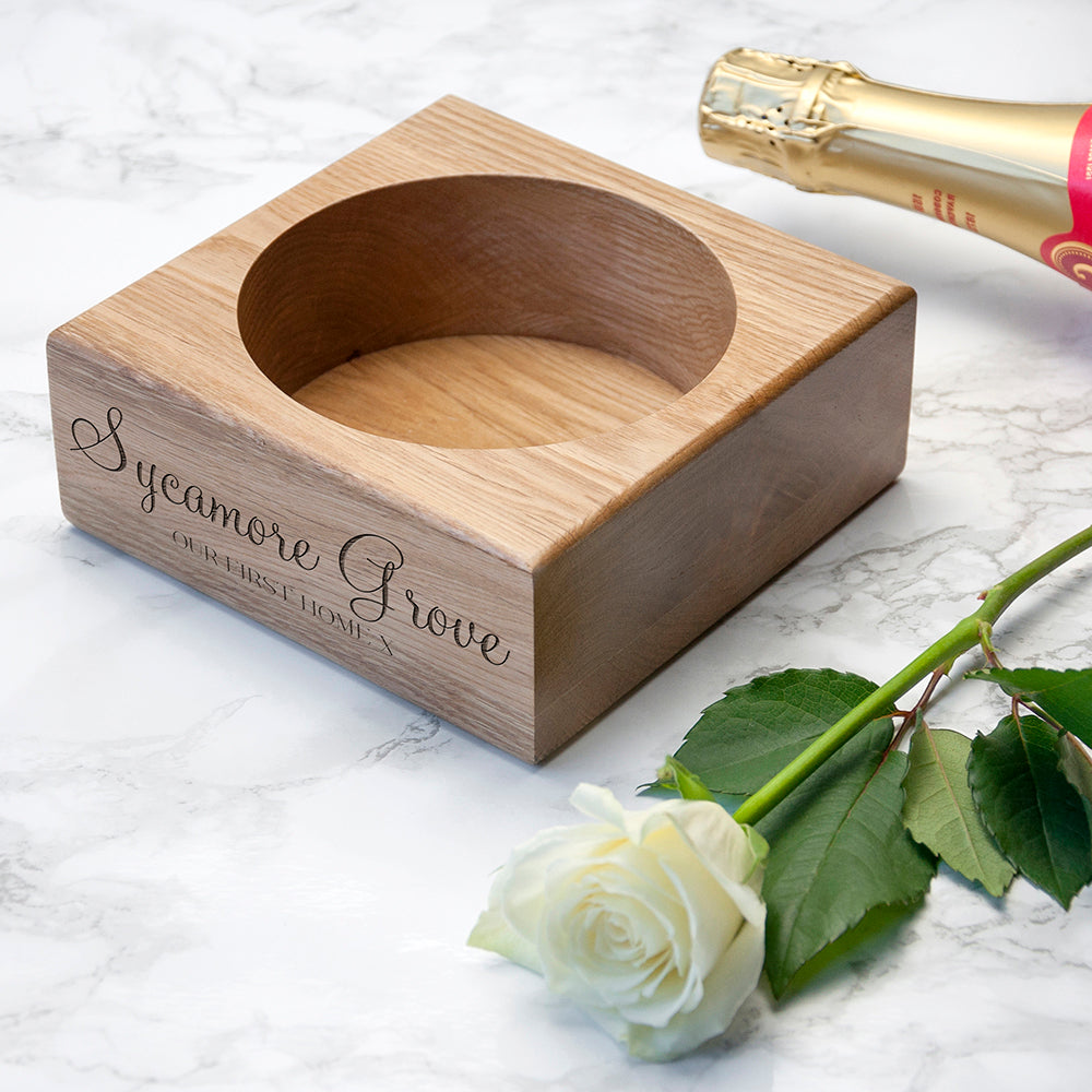 Wooden coaster with customengraving, white rose, and bottle of champagne on a marble surface.