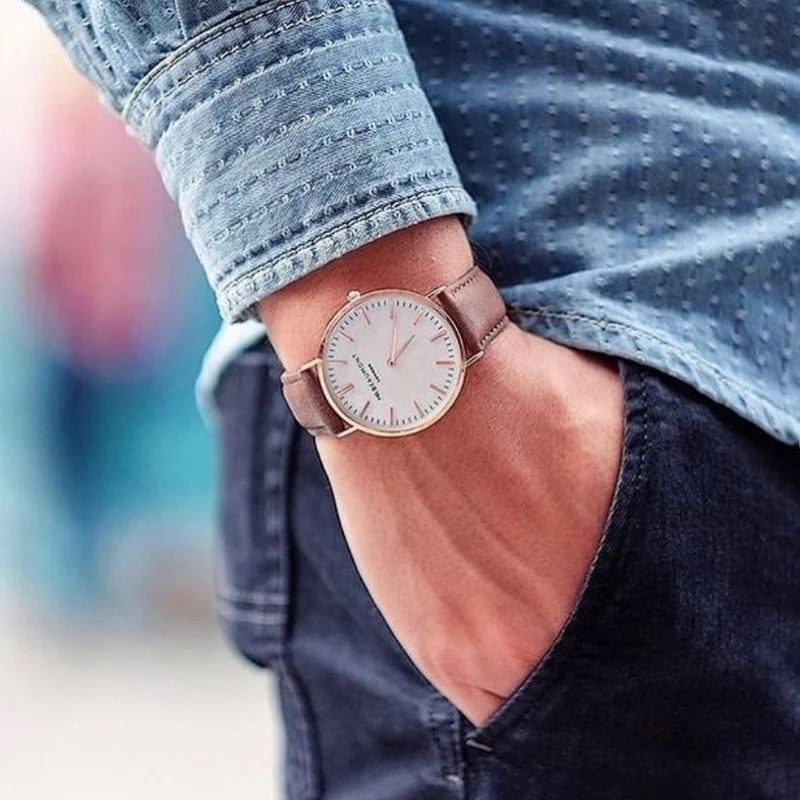 Close-up of a wrist wearing a watch with a brown leather strap and light-colored dial, with denim sleeves and pants.