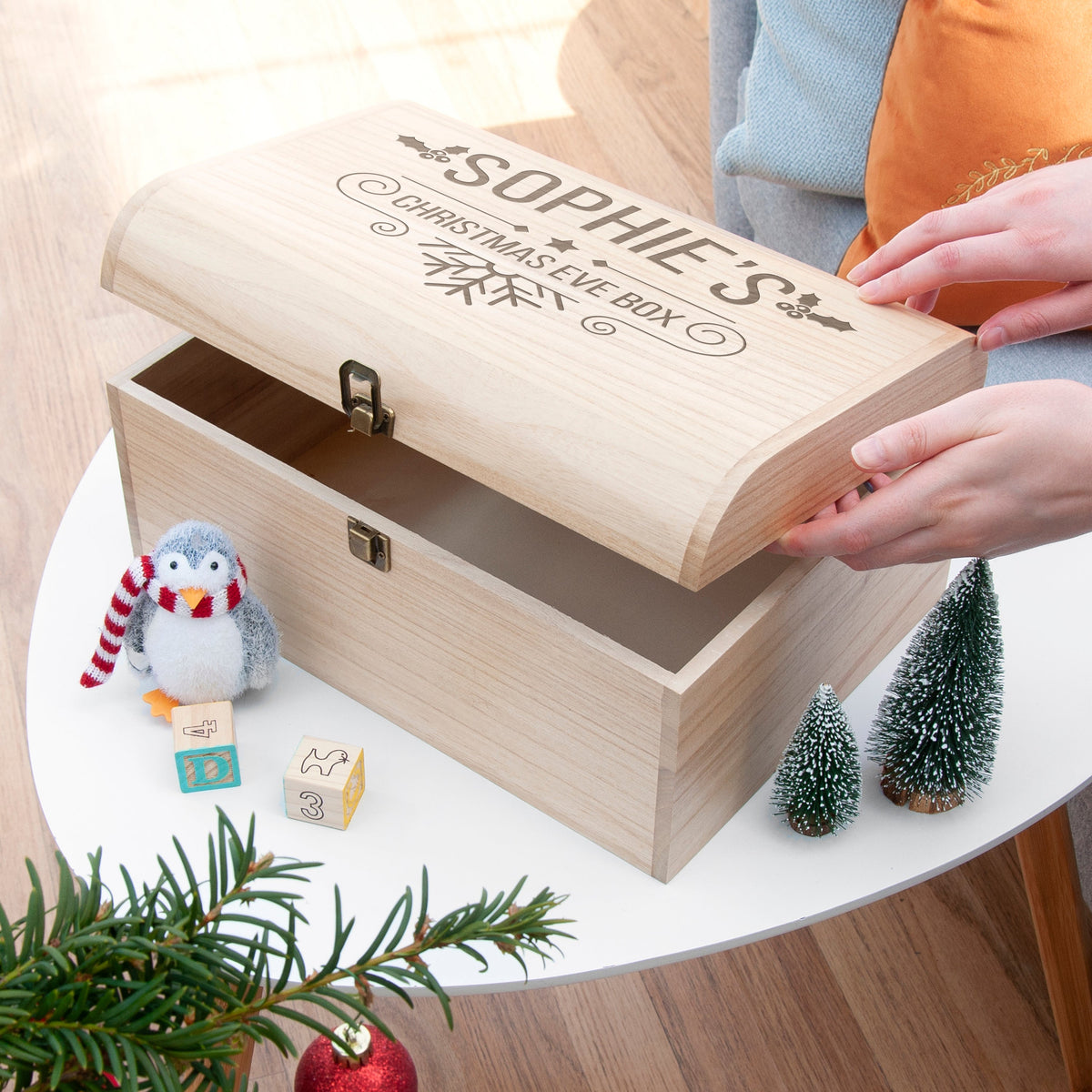 Wooden Christmas Eve box with custom-name engraving, penguin figurine, and small trees on a table.