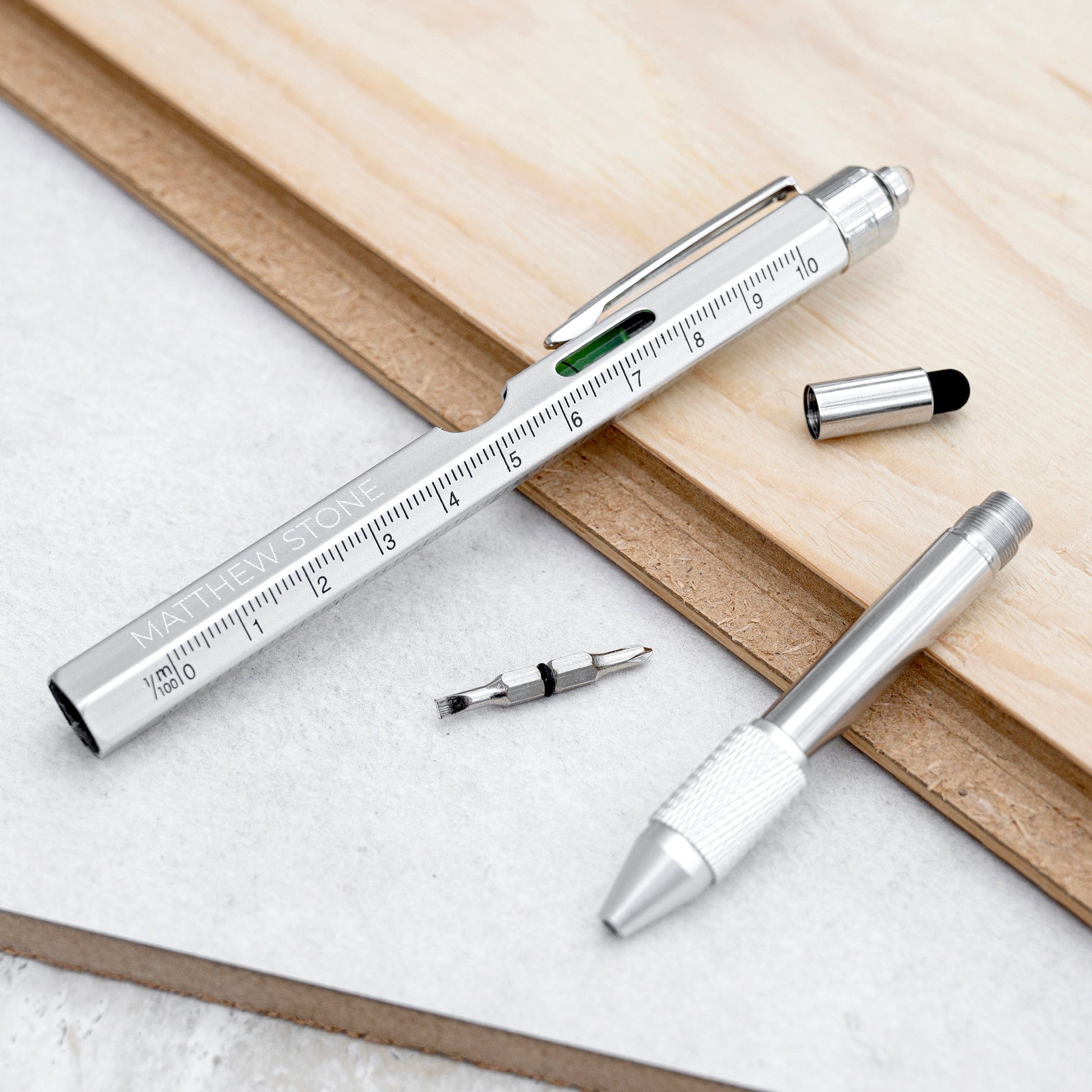 A silver metal pocket utility tool shown disassembled on a light wood and white surface. The pen-like tool has a ruler scale printed on the side, a green window, and small accessories placed nearby.