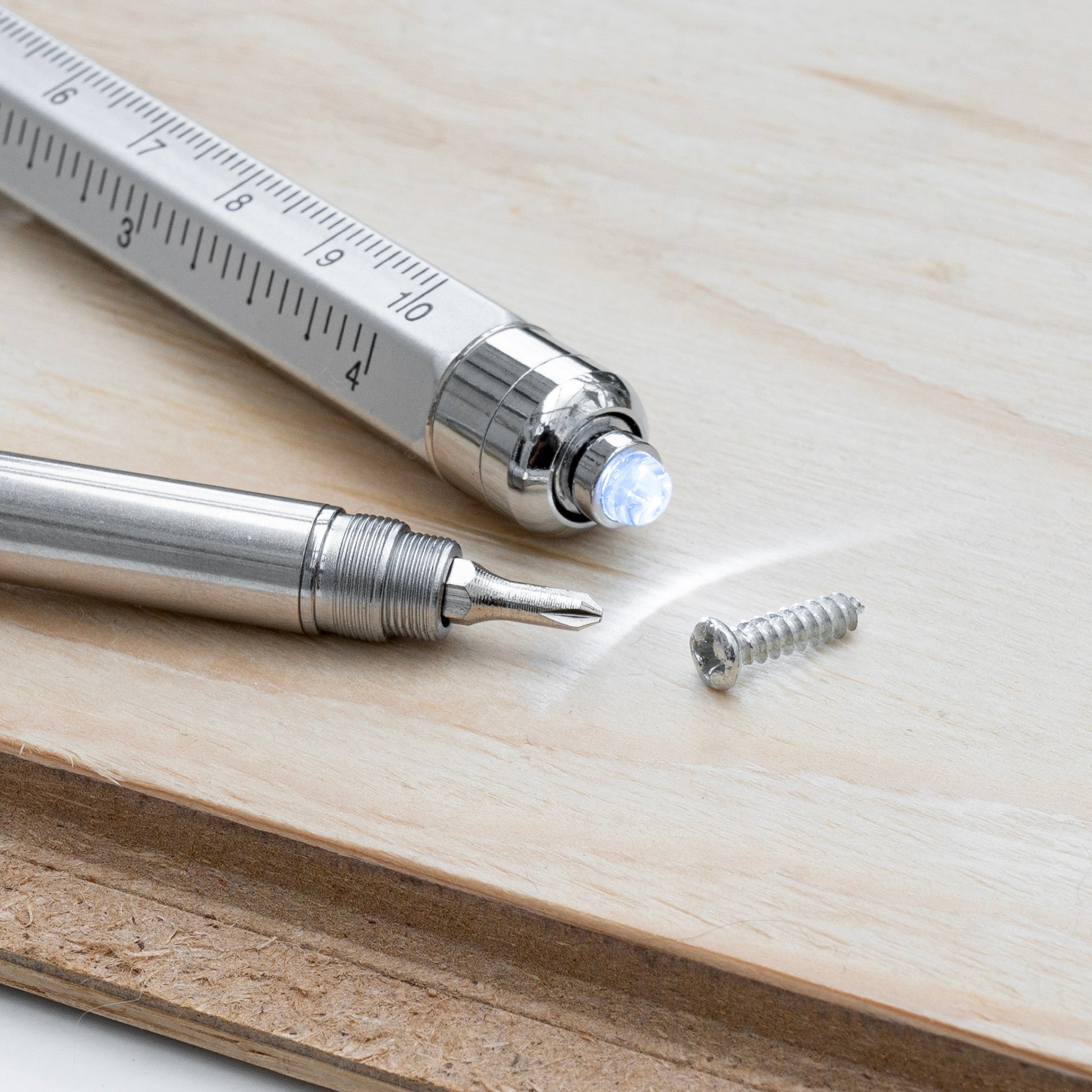 A close-up of a silver metal precision pen tool with a ruler scale and a glowing LED light, shown alongside a separate screw on a light wood surface.