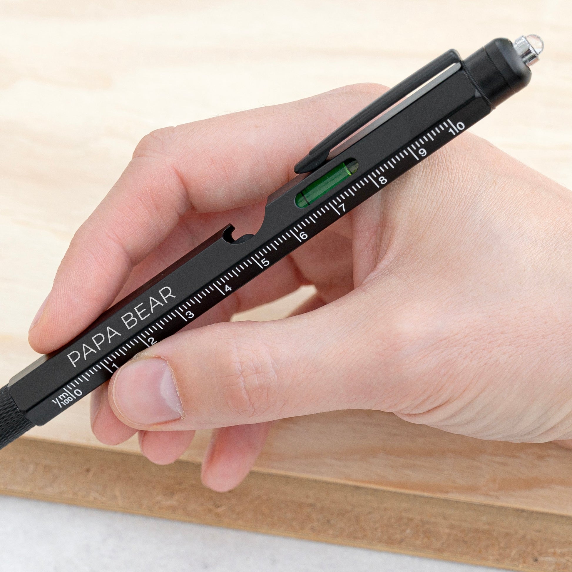 A hand holding a black mechanical pencil with a built-in ruler scale and a green level window. The pencil has 'PAPA BEAR' printed on it, and the background shows a light wood surface.