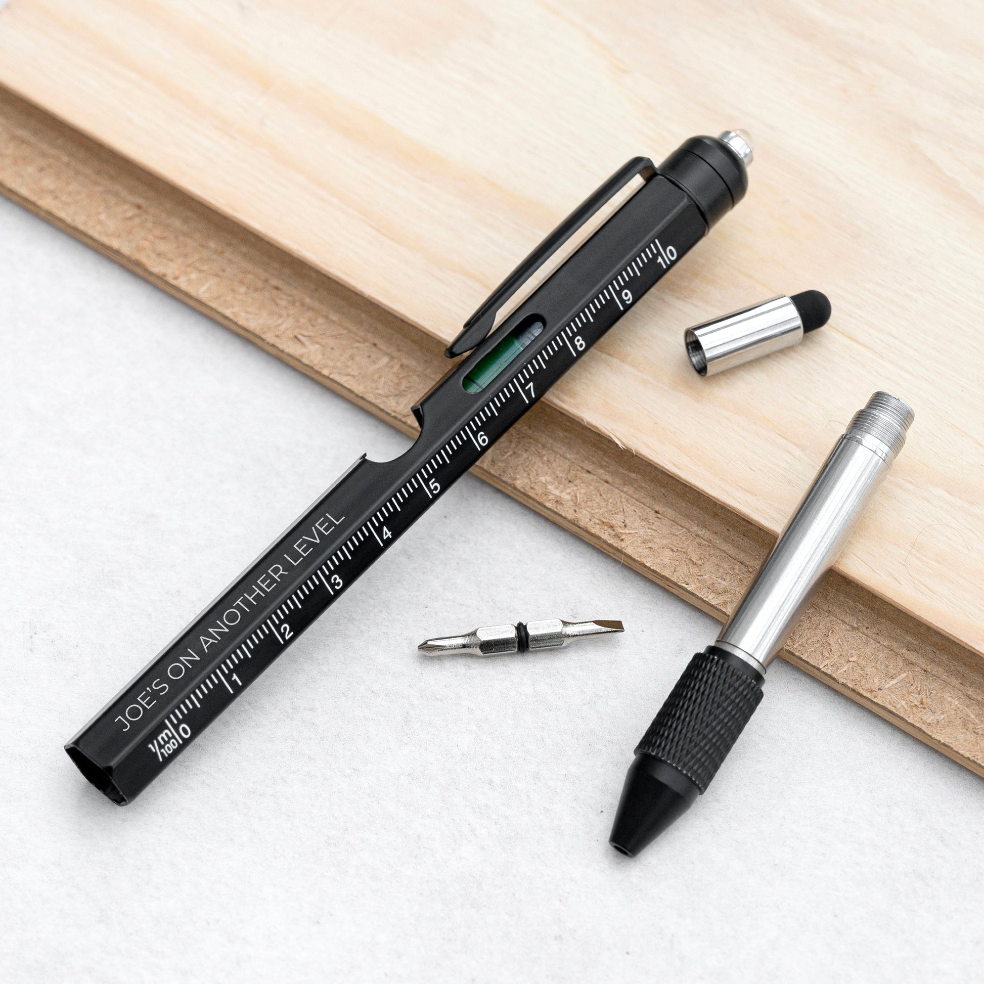 A black and silver multi-tool pocket pen shown with its interchangeable bits on a light wood board and white surface. The pen features a clip, a built-in ruler scale, and a green indicator level.