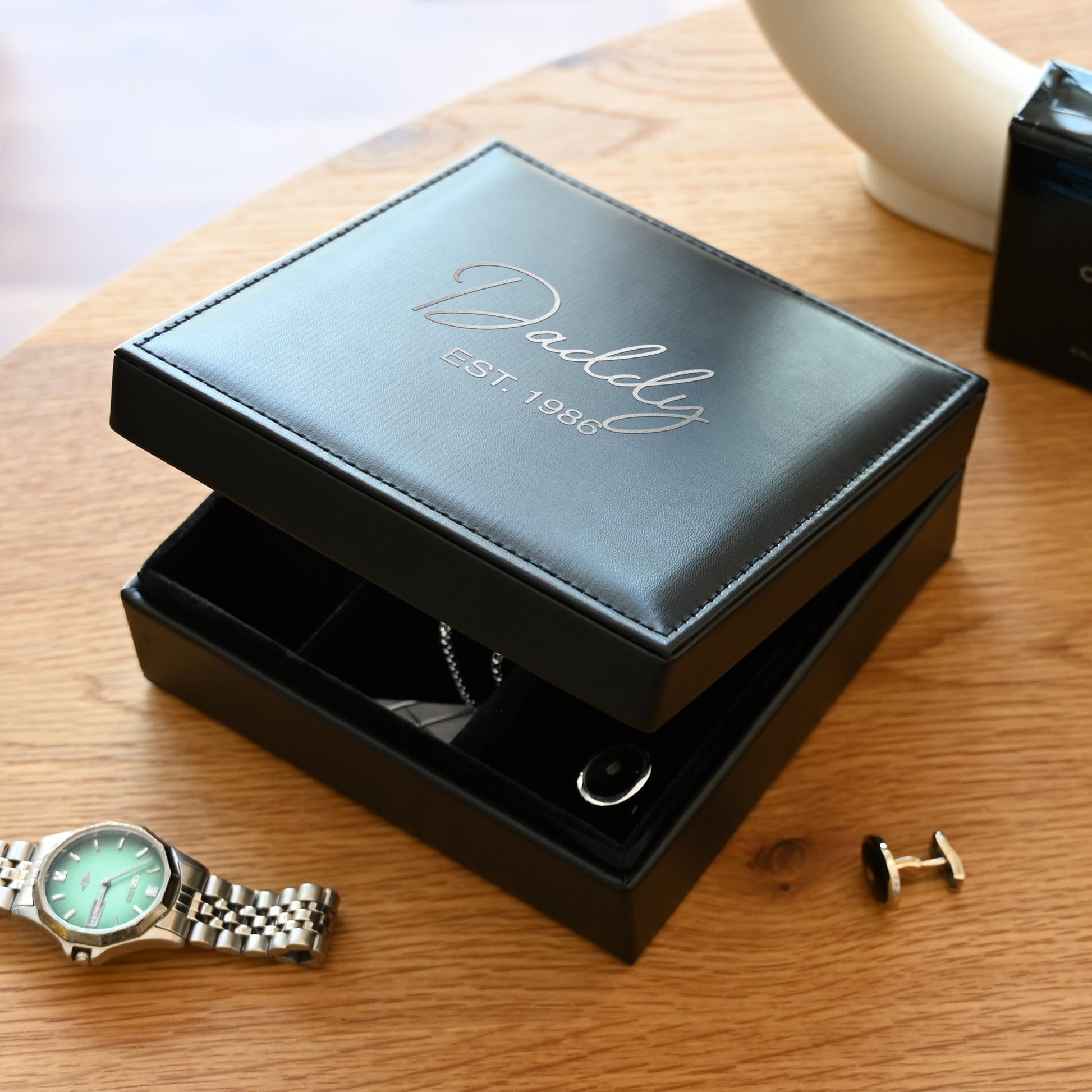 Personalized black leather valet box with custom silver engraving, showing a watch, cufflinks, and jewelry on a wooden table.
