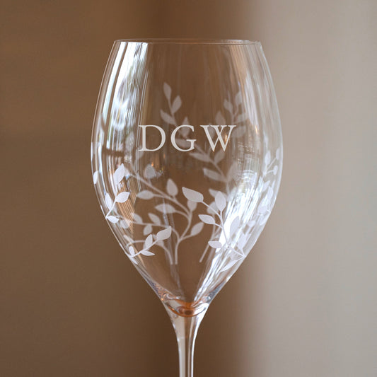 A clear wine glass with etched white floral leaf patterns and the initials 'DGW' on the bowl, photographed against a soft warm beige background.