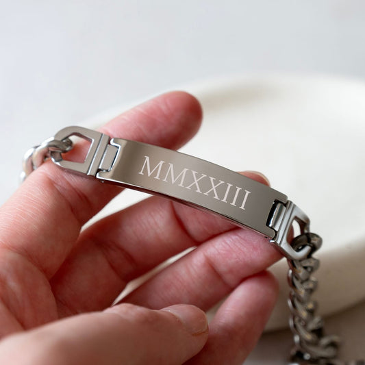 A close-up of a hand holding a silver metal ID bracelet with Roman numerals 'MMXXIII' engraved on the center plate, attached to a chain.