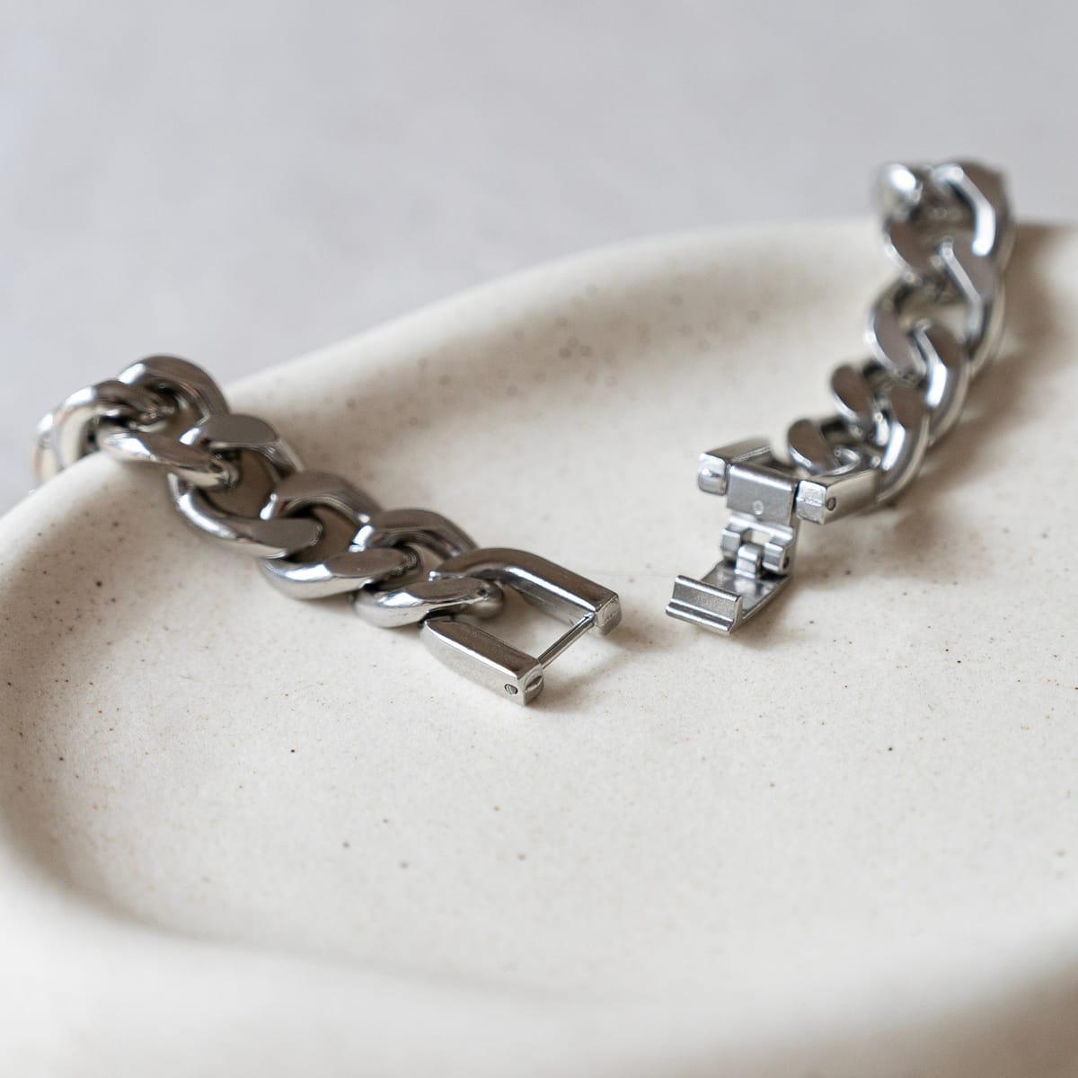 A silver-toned chunky chain bracelet with a unique geometric clasp, displayed on a textured off-white ceramic dish.