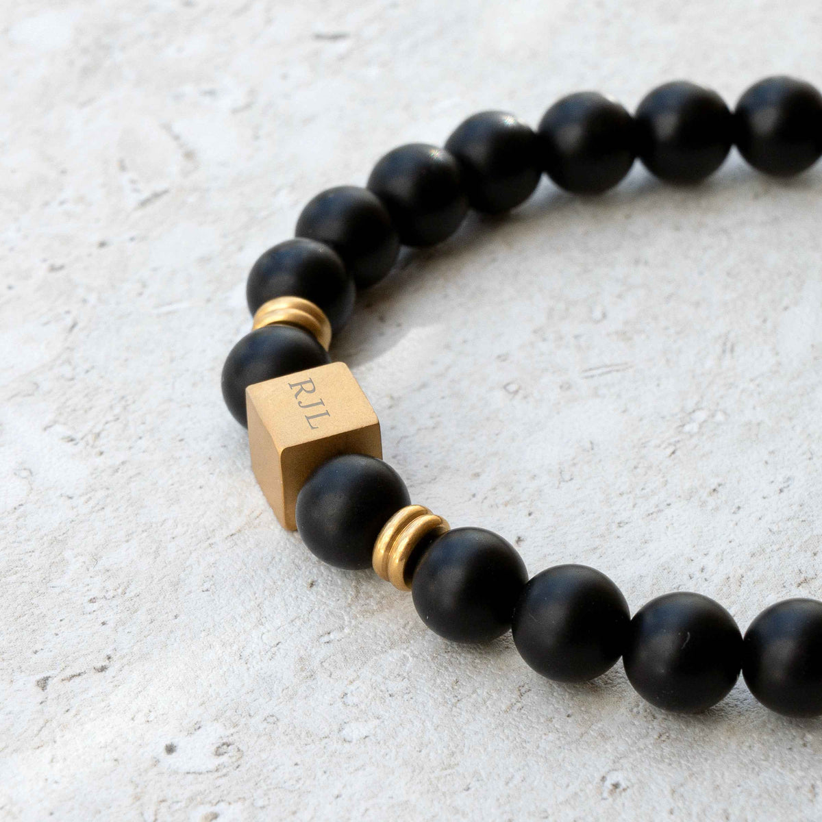 A close-up of a beaded bracelet with matte black round beads, small gold spacer rings, and a square gold charm engraved with 'RJL', shown on a light textured surface.