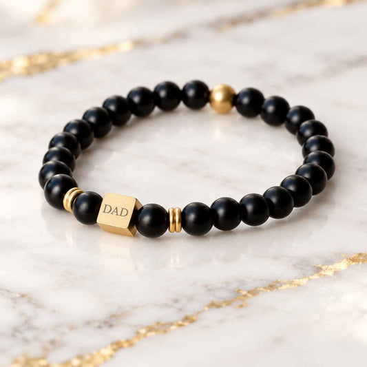 A beaded stretch bracelet with matte black round beads, small gold spacer beads, and a gold cube charm engraved with text