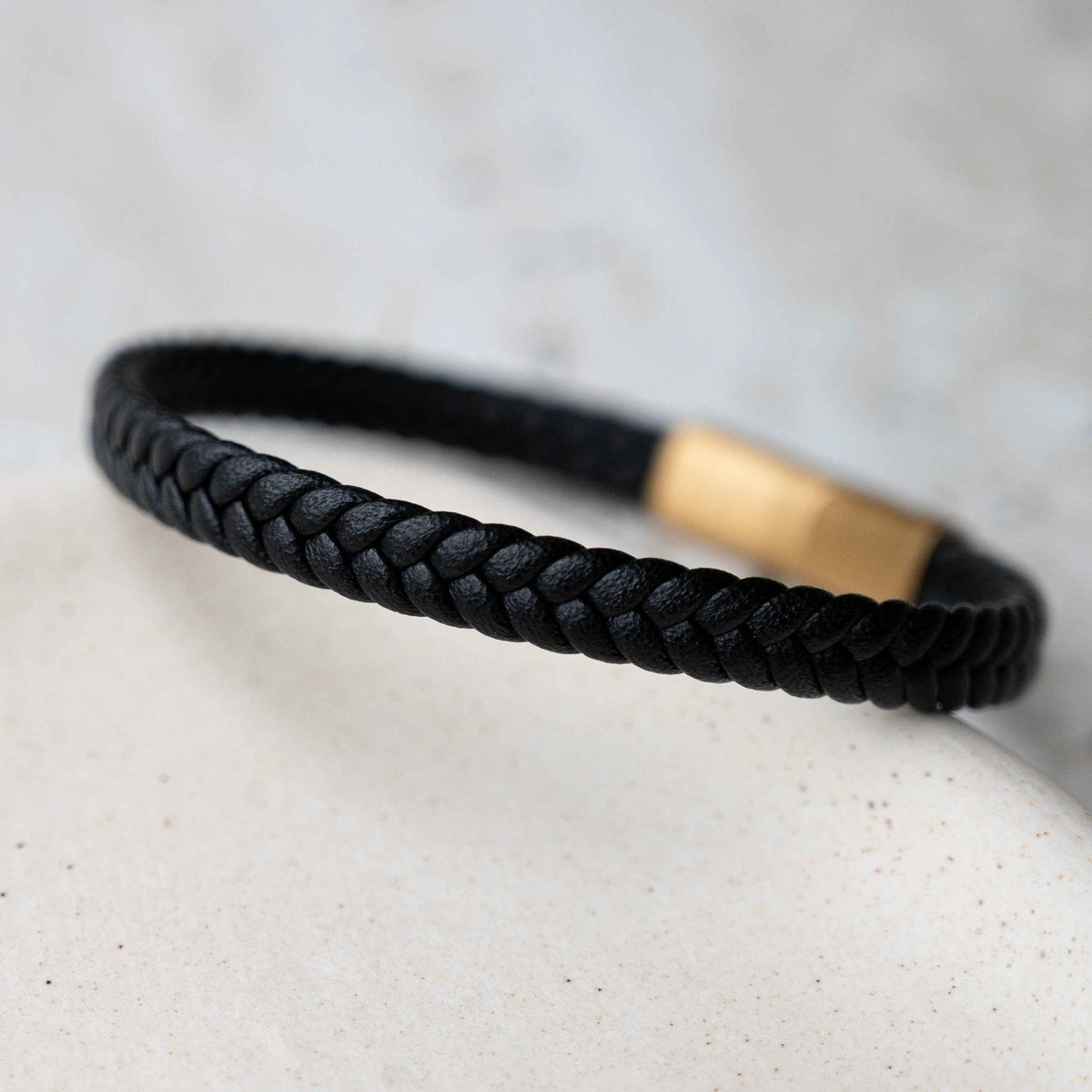 A black braided leather bracelet with a polished gold metal accent, resting on a light beige textured stone against a soft gray background.