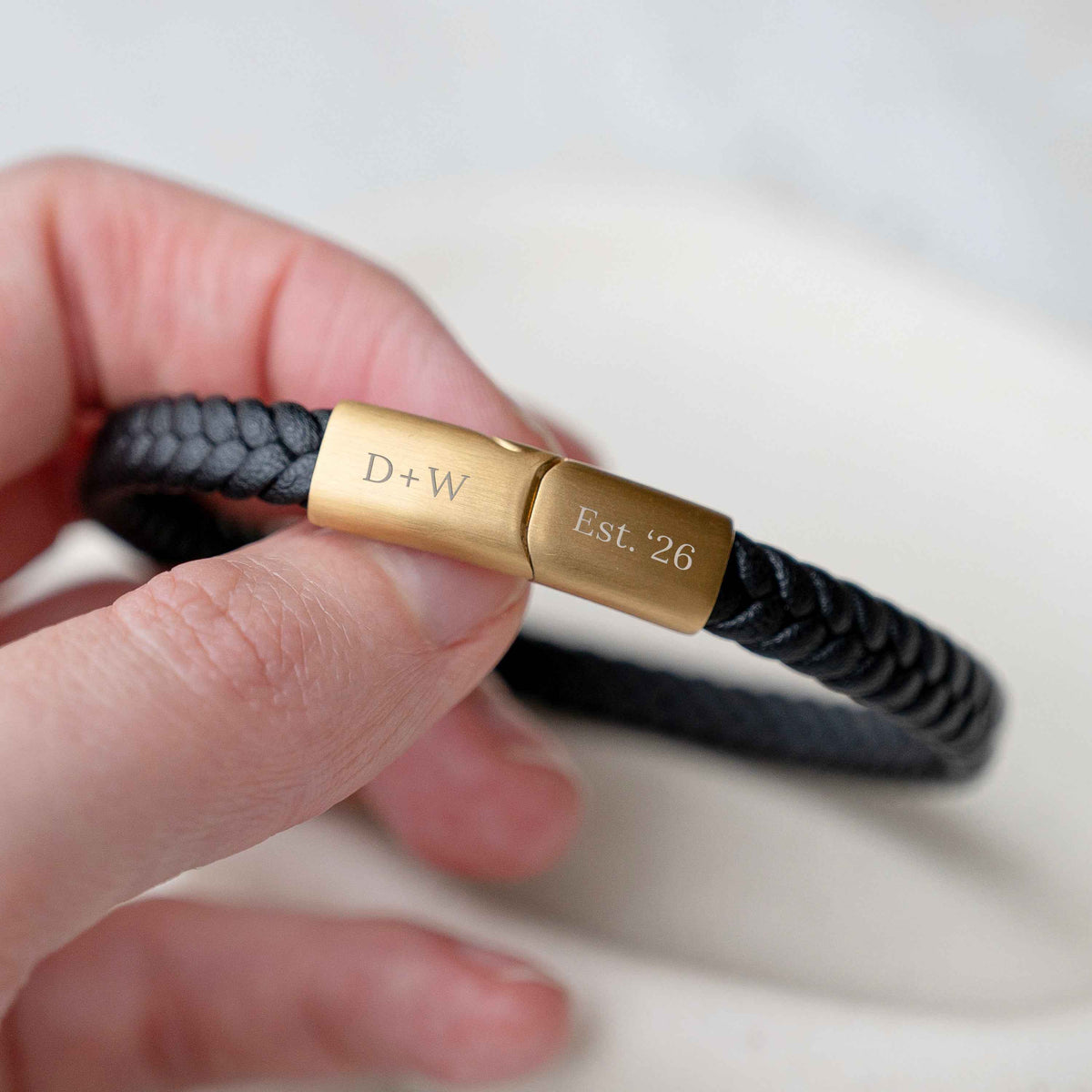 A close-up of a hand holding a black braided leather bracelet with a polished gold-toned clasp engraved with 'D+W' and 'Est. 26'.