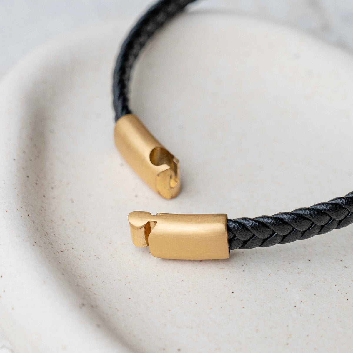 A close-up of a black braided leather bracelet with matte gold metal end caps, resting on a textured off-white ceramic dish.