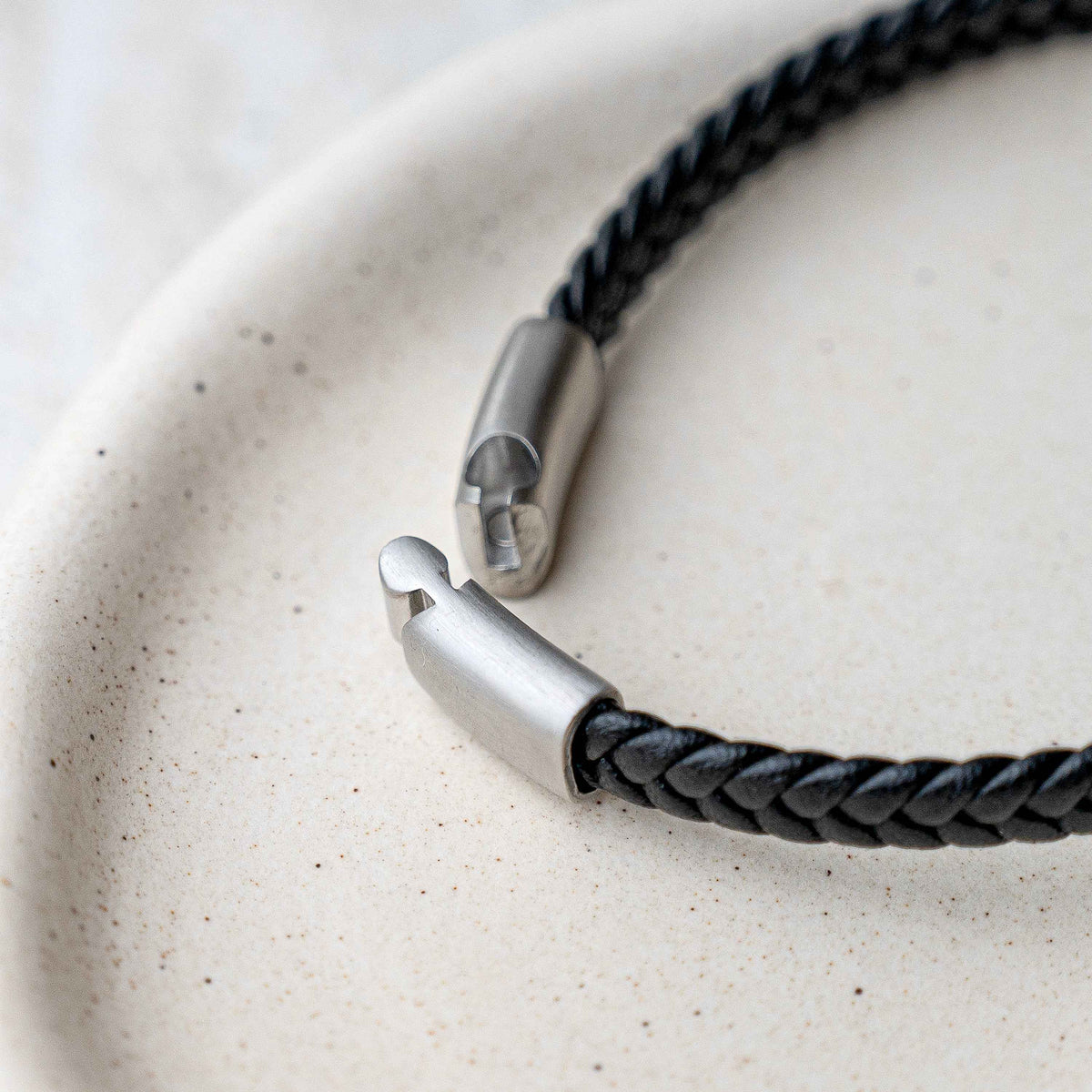 A close-up of a black braided leather bracelet with a silver-toned metal clasp, resting on a textured beige ceramic plate.