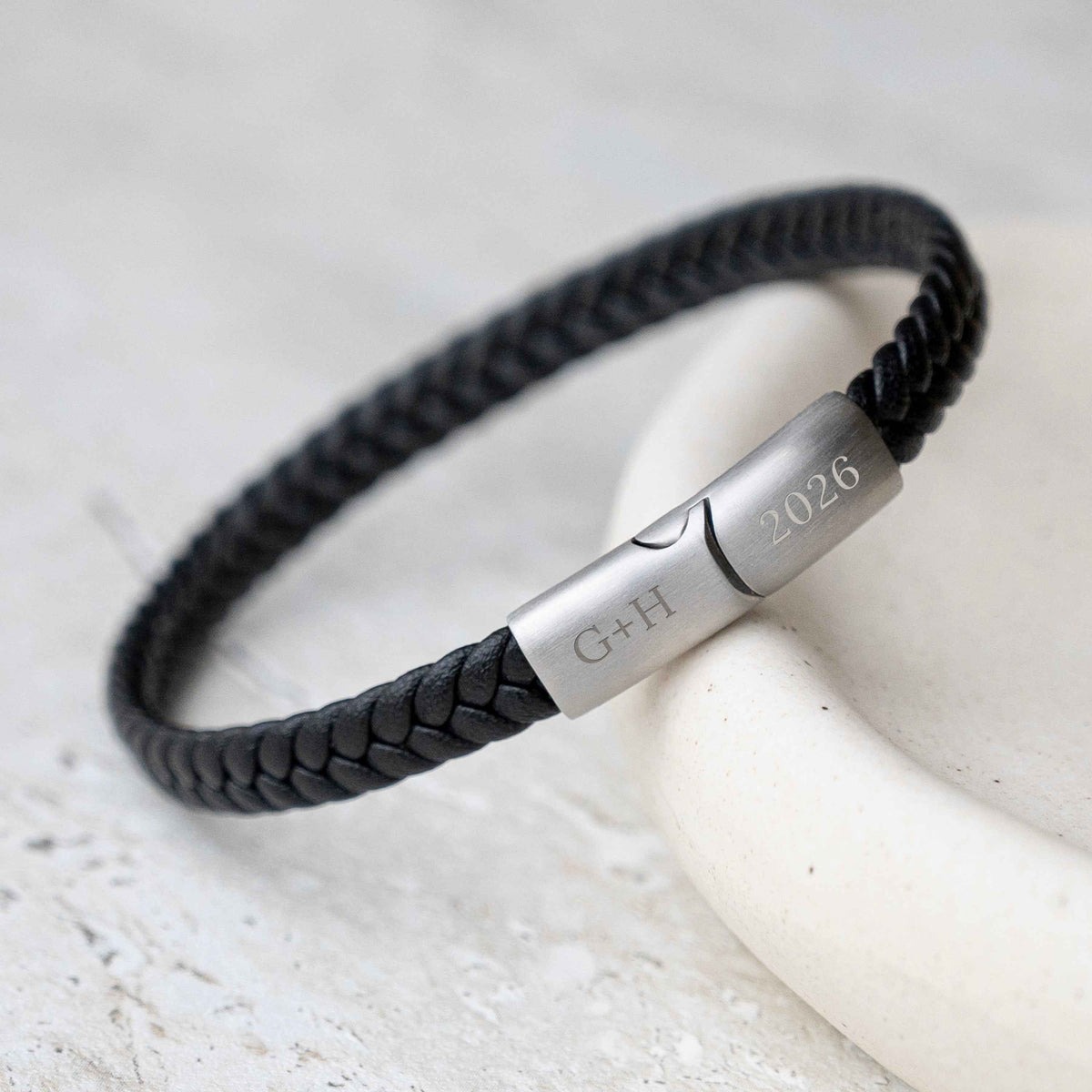 A black braided leather bracelet with a polished silver metal clasp engraved with 'initials and year.