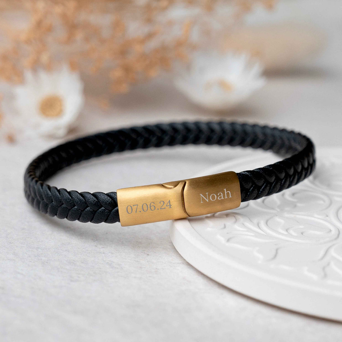 A black braided leather bracelet with a polished gold rectangular clasp engraved with the name 'Noah' and the date '07.06.
