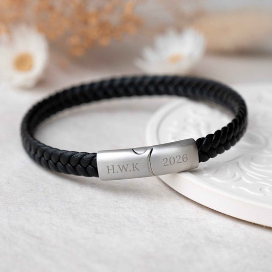 A black braided leather bracelet with a polished silver metal clasp engraved with initials