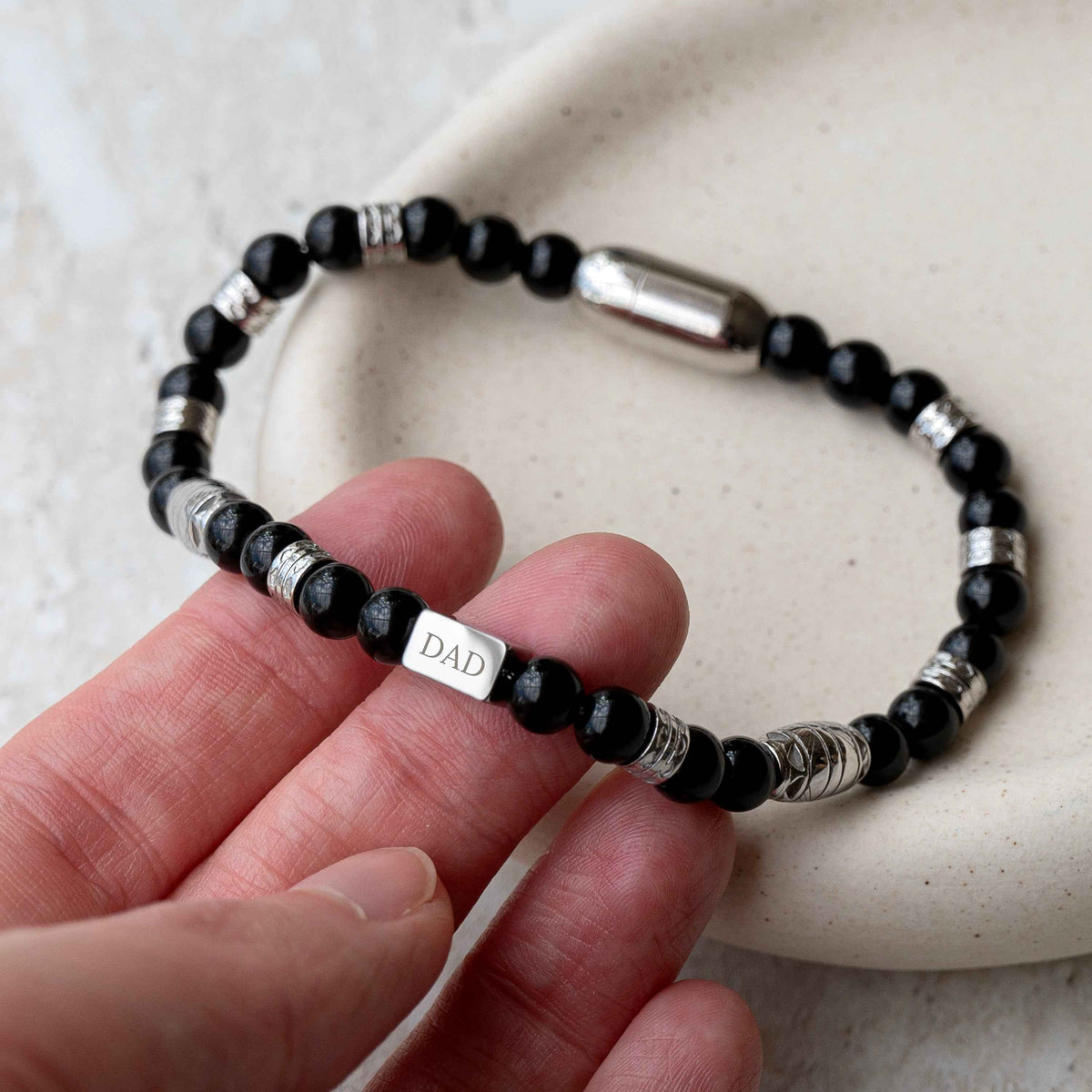 A beaded bracelet with black faceted beads, silver spacer rings, and a central silver cube charm engraved with the word 'DAD'