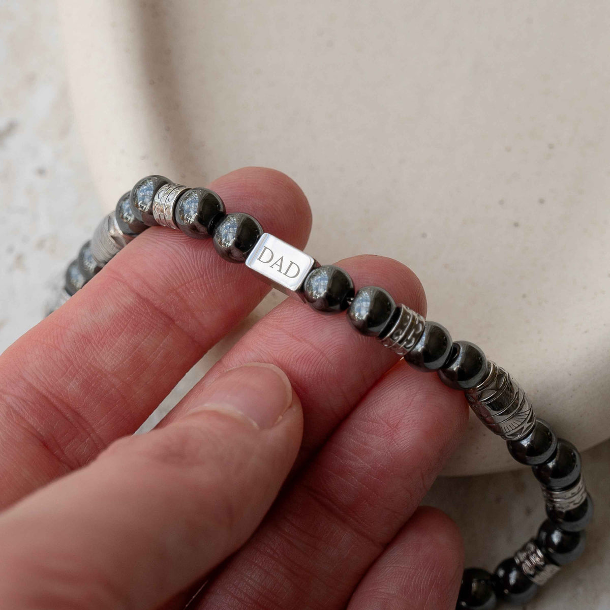 Personalized Hematite-Gemstone Bead Bracelet beaded bracelet, featuring a silver rectangular charm engraved with the word 'DAD'.