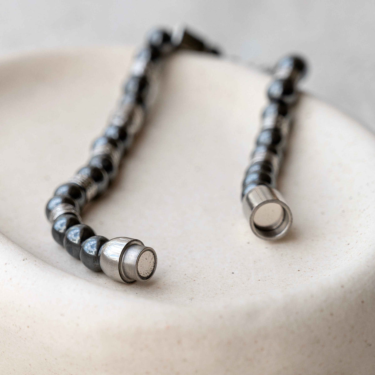 A close-up of a beaded necklace with alternating black and silver metallic beads, resting on a textured beige ceramic surface.