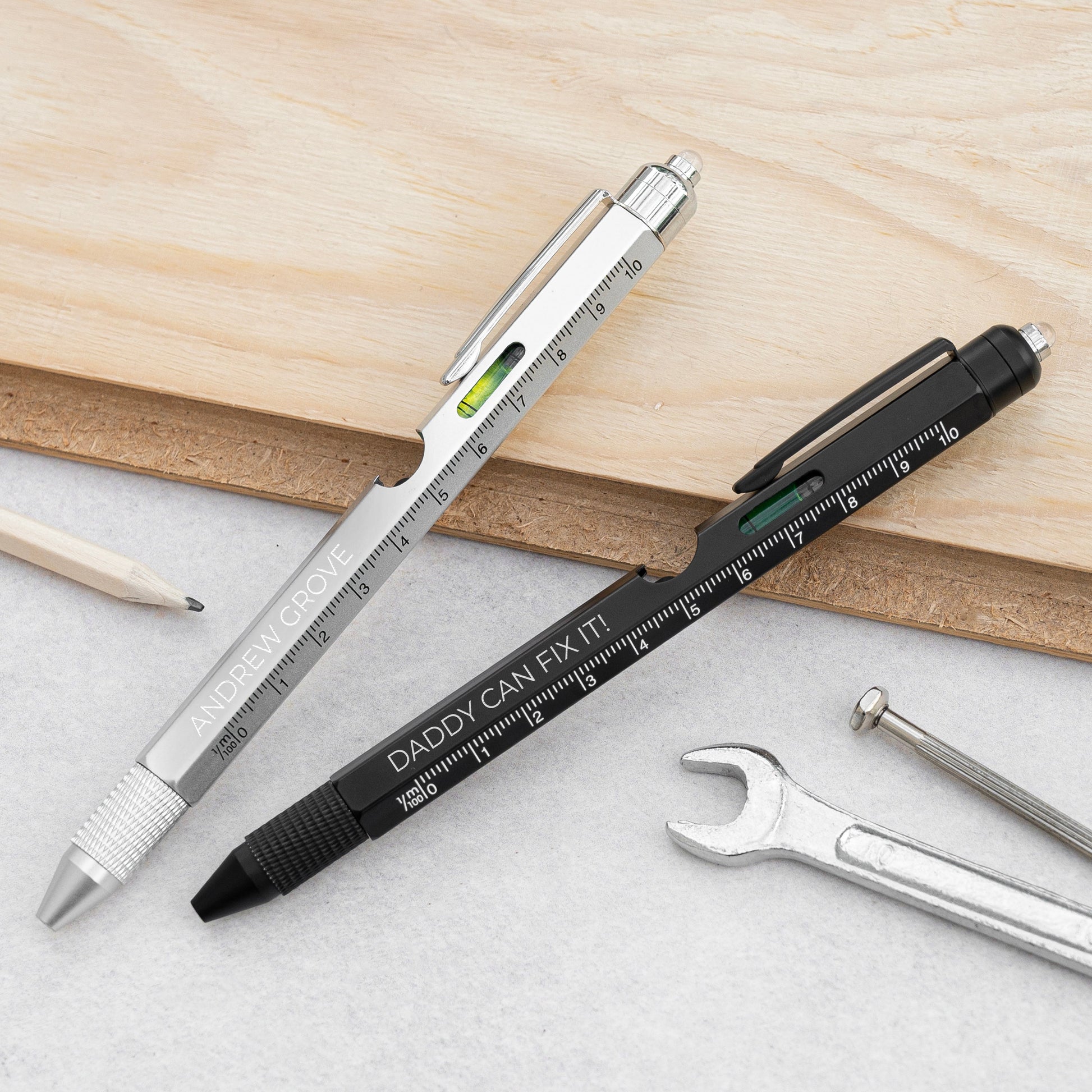 Two multi-tool style pens with ruler markings and small level accents, shown on a light surface with a wooden board, pencil, wrench, and screwdriver nearby. One pen is silver and one is black, both engraved with text including 'DADDY CAN FIX IT!'.