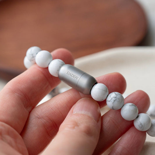 A close-up of a hand holding a beaded bracelet with white and gray marbled beads and a metallic silver capsule in the center engraved with the word 'DADDY'.