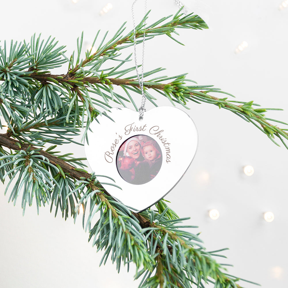Heart-shaped ornament with a photo of a couple and 'Celine's First Christmas' text, hanging on a Christmas tree branch.