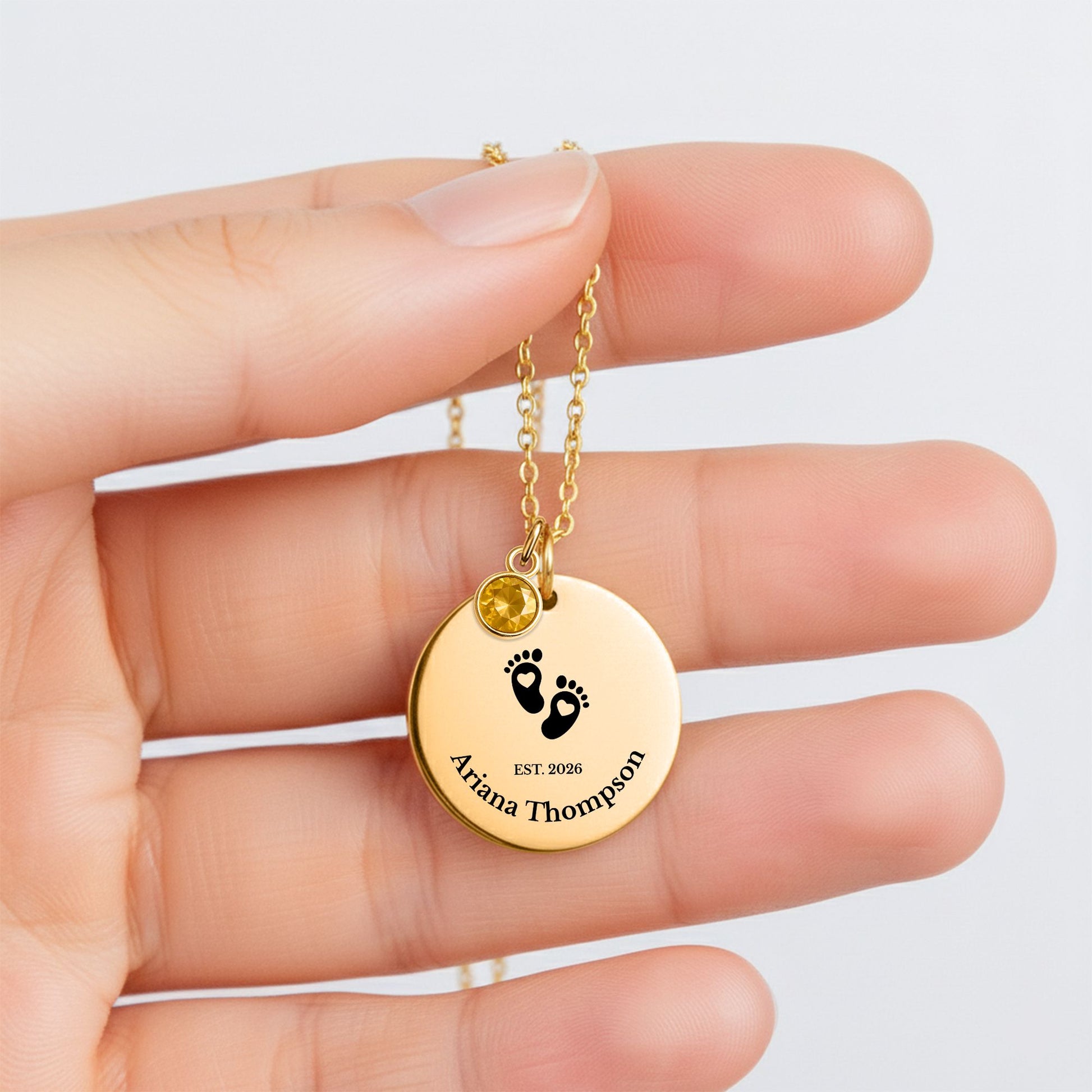 Gold necklace with a round pendant featuring an engraved illustration of a baby, held in a hand against a light background.