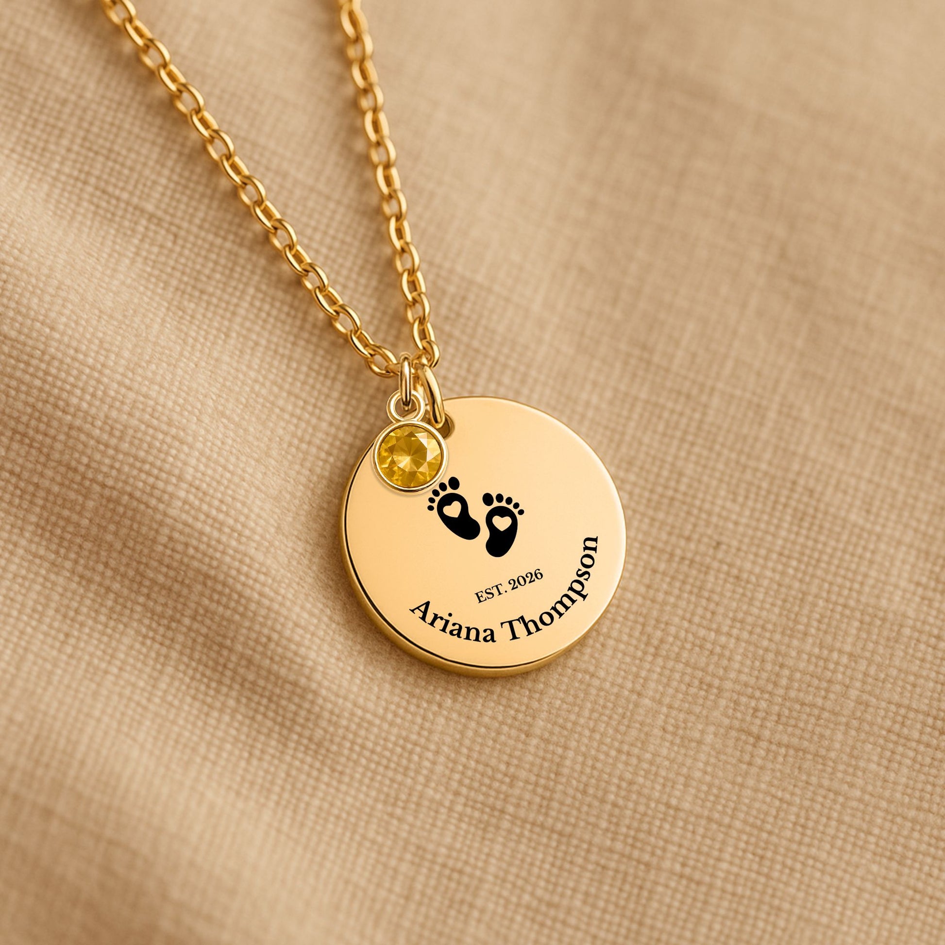A gold personalized necklace with a round disc pendant on a beige textured fabric background. The pendant features black baby footprints, the text 'EST.