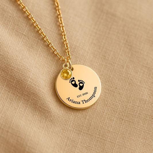 A gold personalized necklace with a round disc pendant on a beige textured fabric background. The pendant features black baby footprints, the text 'EST.