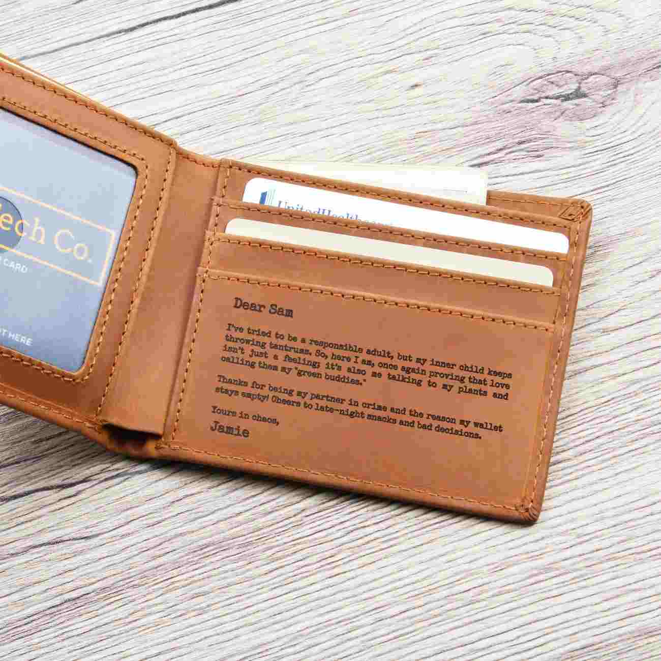 A tan leather bifold wallet shown open on a light wood surface, with card slots, an ID window, and a engraved message pad visible inside.