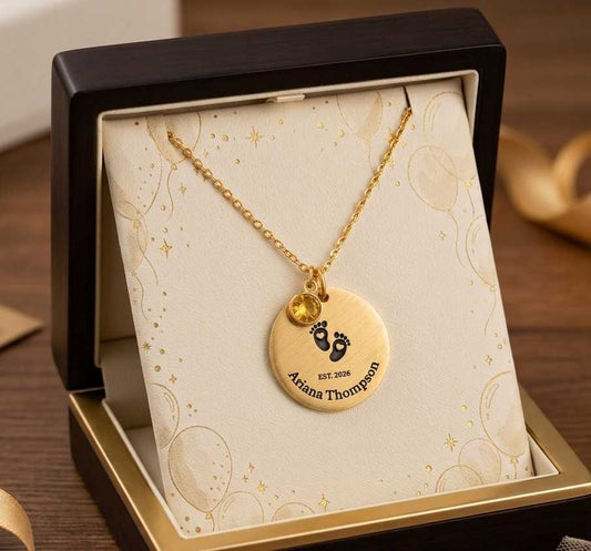 A gold necklace with a round engraved pendant and small yellow gem accents, displayed inside an open black gift box with a cream insert. The pendant is personalized with baby footprints and the name 'Ariana Thompson'.