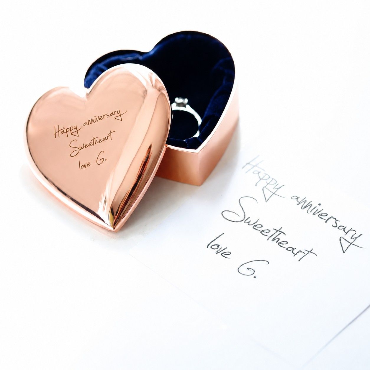 A heart-shaped rose gold keepsake box with one half closed and engraved with 'Happy anniversary Sweetheart love G.', the other half open to reveal a dark blue velvet interior and a ring inside.
