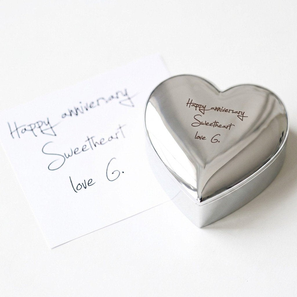 A silver personalized-heart-shaped jewelry box engraved with Happy anniversary message, sits next to a white handwritten note-custom-heart-shaped-jewelry-box-with-actual-handwriting-engraving-silver