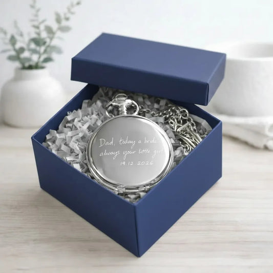 A silver pocket watch pendant engraved with a personalized message, presented inside an open navy blue gift box with shredded white paper on a light wood surface.